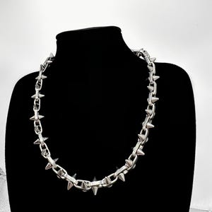 以下が含まれることがあります： 尖ったスパイクが付いたシルバーチェーンネックレス。ネックレスは黒いマネキンに飾られています。