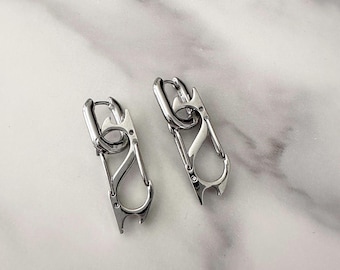 May include: A pair of silver-toned hoop earrings featuring a dangling carabiner-style charm. Each earring consists of a polished metal hoop connected to a functional-looking carabiner clip with a distinct S-shaped curve and a small notch at the base.