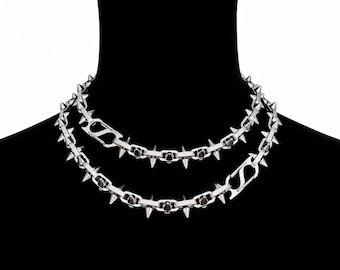 May include: A silver-toned chain necklace featuring a series of uniform metal spikes protruding from each link. The necklace is designed with a double-layered appearance and includes a prominent, stylized S-shaped metal clasp closure.