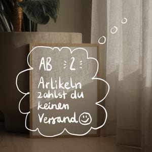 May include: A wooden-framed sign with a cloud-shaped border displays handwritten text in German. The text reads "AB 2: Artikeln zahlst du keinen Versand" with a smiley face. The sign is on a wooden floor, near a potted plant and sheer curtains.