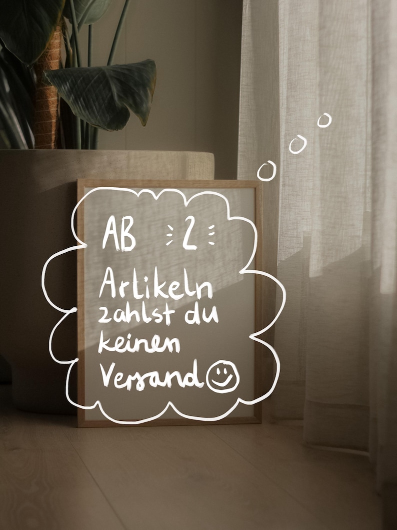 May include: A wooden-framed sign with a cloud-shaped border displays handwritten text in German. The text reads "AB 2: Artikeln zahlst du keinen Versand" with a smiley face. The sign is on a wooden floor, near a potted plant and sheer curtains.