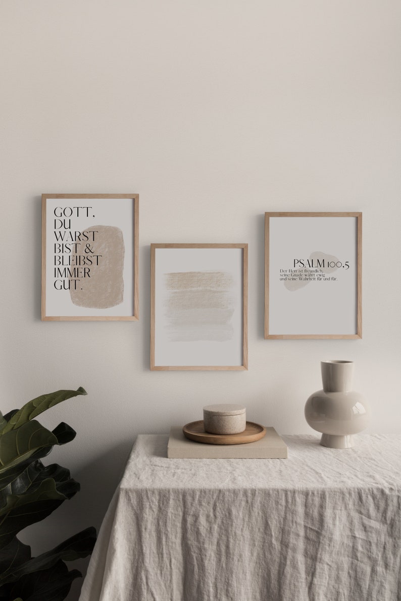 May include: Three framed prints on a white wall above a light brown table with a vase and a small box. The prints are all in a light wood frame. The left print has a beige background with the text "Gott. Du warst bist & bleibst immer gut." The middle print has a beige abstract brushstroke design. The right print has a beige background with the text "Psalm 100.5 Der Herr ist freundlich, seine Gnade w&auml;hrt ewig, und seine Wahrheit f&uuml;r und f&uuml;r."