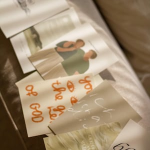 May include: A collection of folded cards with various designs and text. Some cards feature illustrations of people, while others have handwritten phrases in orange and white. The cards are arranged on a soft, neutral-toned surface.