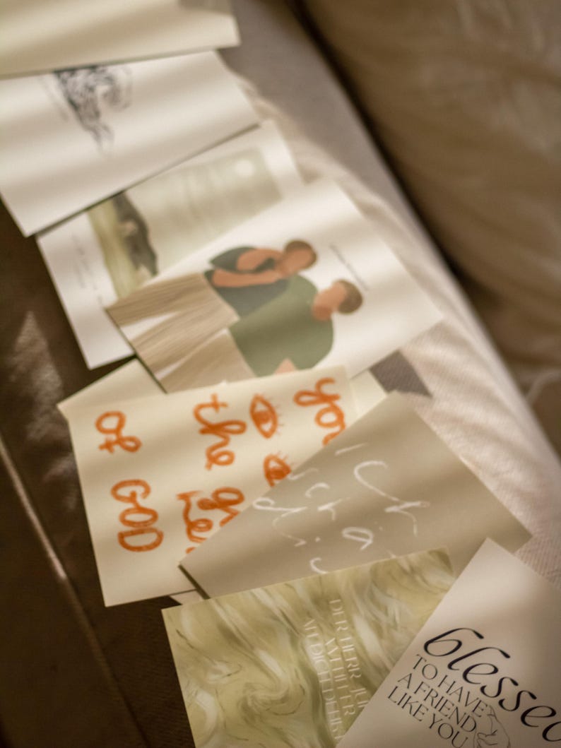 May include: A collection of folded cards with various designs and text. Some cards feature illustrations of people, while others have handwritten phrases in orange and white. The cards are arranged on a soft, neutral-toned surface.