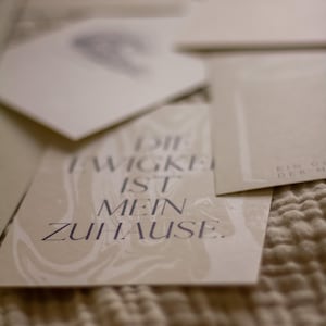 May include: A collection of off-white cards with text in a dark purple font. The cards are arranged on a textured, beige surface. One card reads "DIE EWIGKEIT IST MEIN ZUHAUSE."