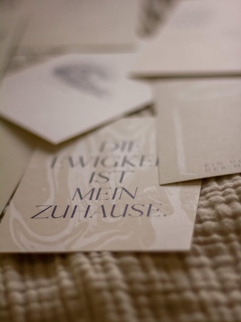 May include: A collection of off-white cards with text in a dark purple font. The cards are arranged on a textured, beige surface. One card reads "DIE EWIGKEIT IST MEIN ZUHAUSE."