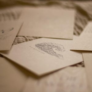 May include: A collection of cream-colored stationery cards with delicate gray illustrations. One card features a wave design, while others have floral or abstract patterns. The cards are arranged on a textured surface.