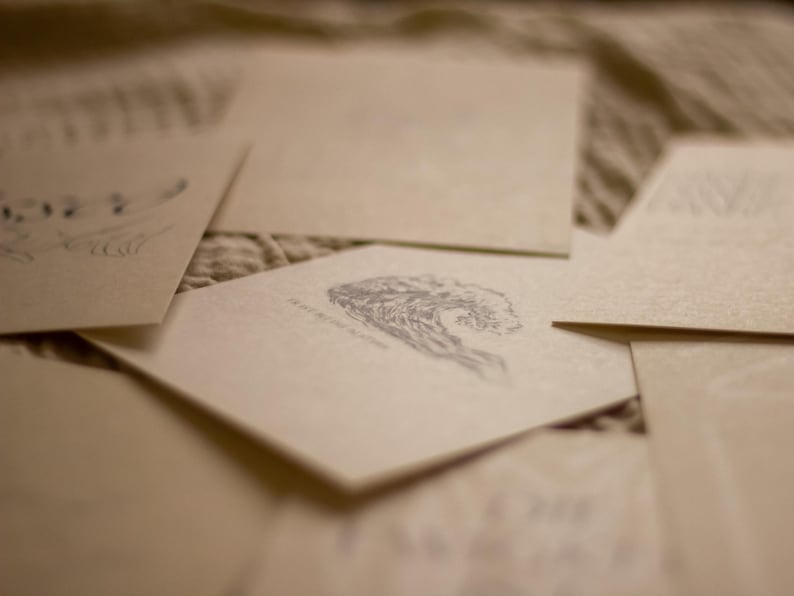 May include: A collection of cream-colored stationery cards with delicate gray illustrations. One card features a wave design, while others have floral or abstract patterns. The cards are arranged on a textured surface.