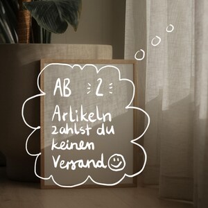 May include: A framed sign with a white handwritten message that says "AB 2: Artikeln zahlst du keinen Versand" with a smiley face. The sign is on a wooden floor in front of a white curtain.