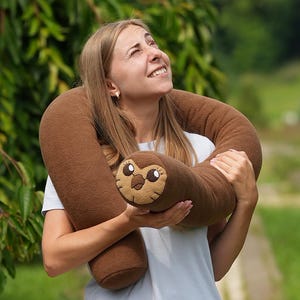Puede incluir: Una almohada de felpa con forma de perezoso marrón con una cara sonriente es sostenida por una persona. La felpa tiene un cuerpo largo y cilíndrico y una cabeza redonda con rasgos faciales bordados. El fondo es un entorno verde al aire libre.