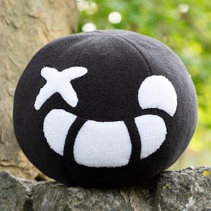 May include: A black plush toy shaped like a sphere with a white smiling face. The face has a white X for an eye and a white crescent moon shape for a mouth.