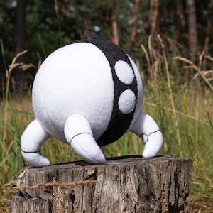 Peut inclure: Un jouet en peluche blanc et noir avec trois pattes et un corps rond. Le jouet est assis sur une souche d'arbre dans un champ herbeux.