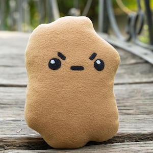 May include: A brown plush toy shaped like a potato with a grumpy expression. The potato has black eyes, a black mouth, and black eyebrows.