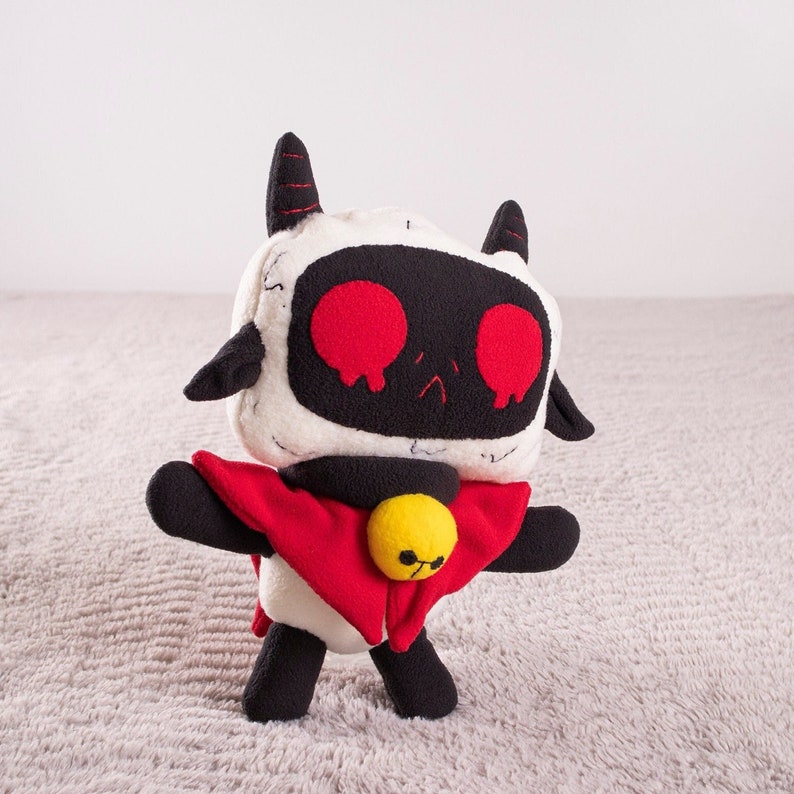 May include: A plush toy robot with a white body, black head, and red eyes. The robot is wearing a red cape and has a yellow bell on its chest.
