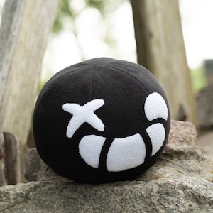 Puede incluir: Pelota de felpa negra con una cara sonriente cosida en blanco. La cara tiene una cruz para un ojo y una l&iacute;nea curva para una boca.