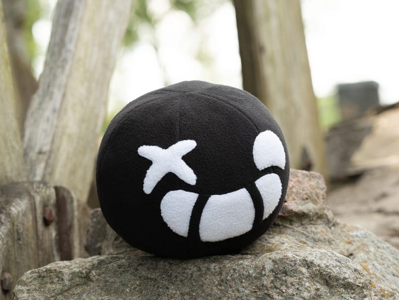 Puede incluir: Pelota de felpa negra con una cara sonriente cosida en blanco. La cara tiene una cruz para un ojo y una l&iacute;nea curva para una boca.