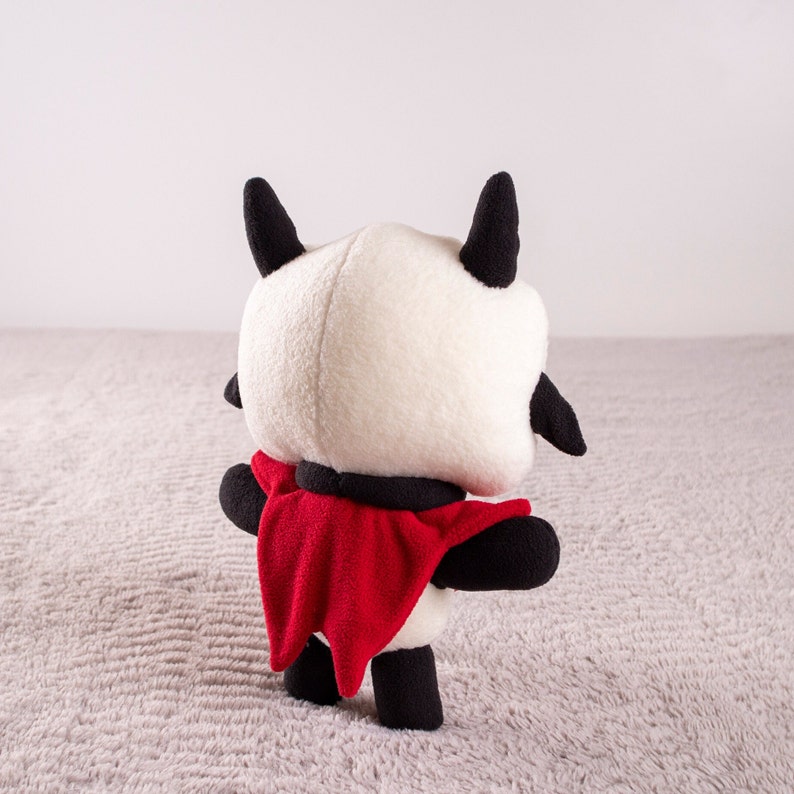 May include: A plush toy with a white body, black horns, and a red cape. The toy is standing on a light gray surface.