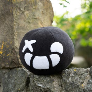 Puede incluir: Pelota de felpa negra con una cara sonriente blanca y una X por un ojo. La pelota est&aacute; sentada sobre una roca.