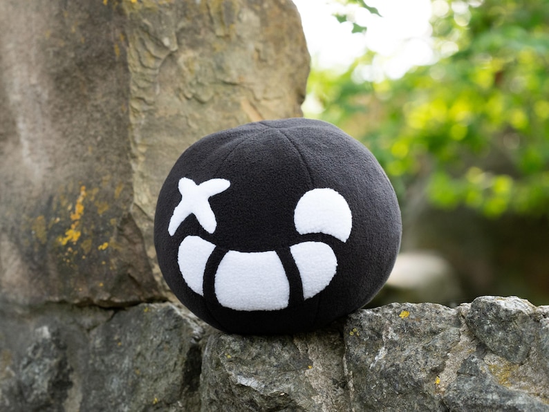 Puede incluir: Pelota de felpa negra con una cara sonriente blanca y una X por un ojo. La pelota est&aacute; sentada sobre una roca.