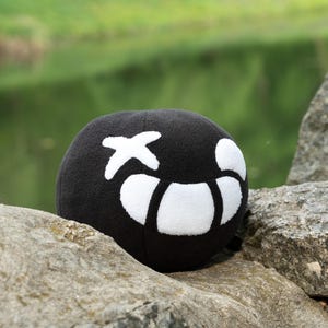Puede incluir: Un juguete de peluche negro con forma de bola con una X blanca y un dise&ntilde;o de media luna blanca. El juguete est&aacute; sentado en una roca.