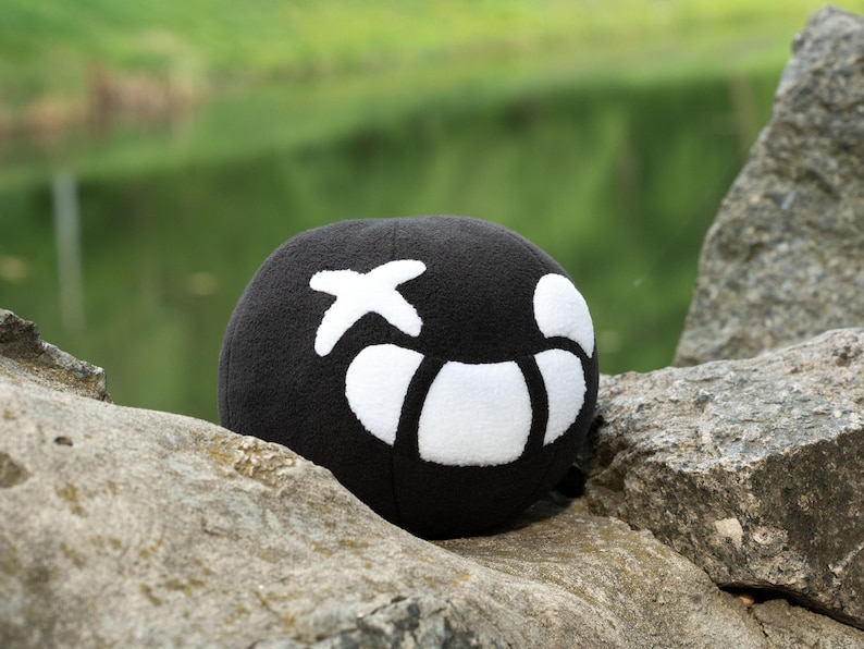 Puede incluir: Un juguete de peluche negro con forma de bola con una X blanca y un dise&ntilde;o de media luna blanca. El juguete est&aacute; sentado en una roca.
