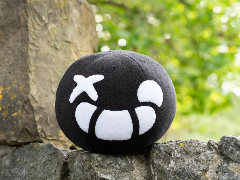 Puede incluir: Un juguete de peluche negro con forma de esfera con una cara sonriente blanca. La cara tiene una X blanca para un ojo y una forma de media luna blanca para una boca.