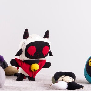 May include: A group of five plush toys, including a white and black robot-like toy with red eyes and a red cape, a black and gray toy with a swirl design, a blue toy with yellow buttons, and two other toys with a patchwork design.