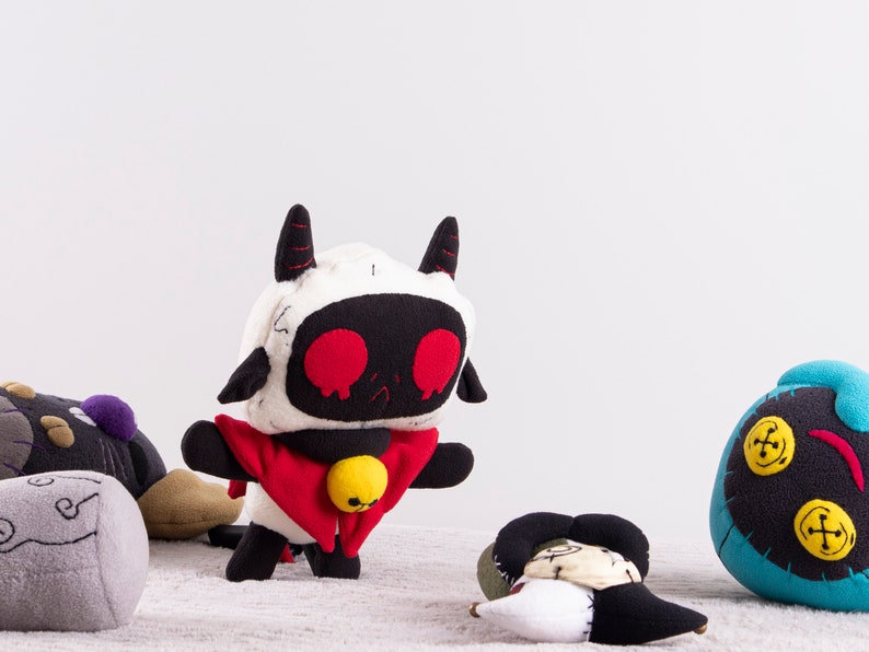 May include: A group of five plush toys, including a white and black robot-like toy with red eyes and a red cape, a black and gray toy with a swirl design, a blue toy with yellow buttons, and two other toys with a patchwork design.