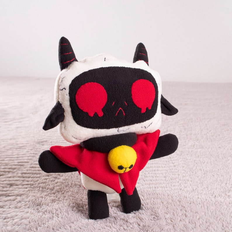 May include: A white and black plush toy with red horns and a red cape. The toy has large red eyes and a yellow bell on its chest.