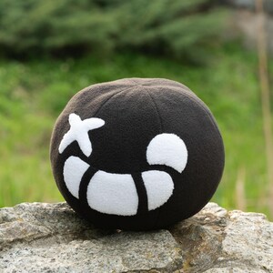 Puede incluir: Un juguete de peluche negro con forma de calavera con un dise&ntilde;o de cruz blanca y media luna blanca. El juguete est&aacute; sentado en una roca.