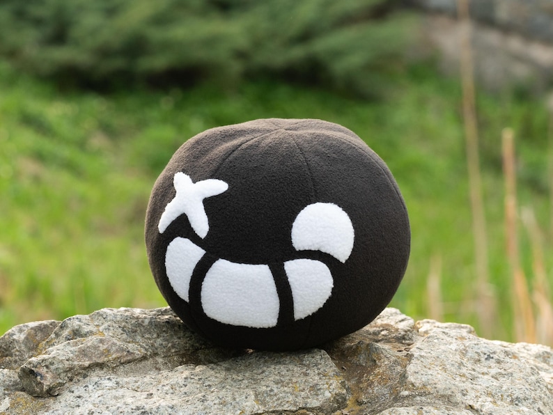 Puede incluir: Un juguete de peluche negro con forma de calavera con un dise&ntilde;o de cruz blanca y media luna blanca. El juguete est&aacute; sentado en una roca.