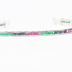 May include: A silver bracelet featuring a row of emerald, ruby, and sapphire gemstones.
