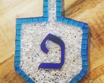 May include: A dreidel ornament featuring a mosaic border of small blue glass tiles and a center filled with crushed clear glass fragments. A dark blue Hebrew letter Peh is centered on the surface, and a vertical handle extends from the top.