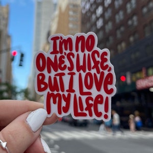 Puede incluir: Una pegatina blanca con letras rojas que dice "No soy la esposa de nadie, pero amo mi vida!" La pegatina se sostiene en una mano contra un fondo borroso de la ciudad.