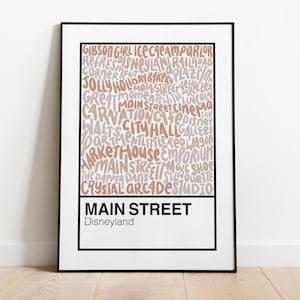May include: A framed print with a white background and black frame. The print features a word cloud of various shops and attractions found on Main Street, U.S.A. at Disneyland. The words are in a light brown and gray color scheme. The text at the bottom of the print reads "Main Street, Disneyland."