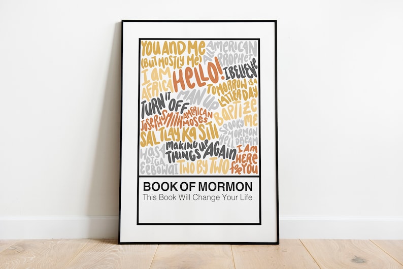 May include: A framed poster with a white background and black frame. The poster features a word cloud of text in various shades of yellow, orange, and gray. The text includes phrases like "You and Me", "Hello", "Turn it Off", "Book of Mormon", and "This Book Will Change Your Life".