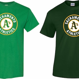 Peut inclure: Deux t-shirts, un vert et un vert foncé, avec un logo circulaire blanc comportant le texte "Sacramento A's Athletics" en lettres dorées.