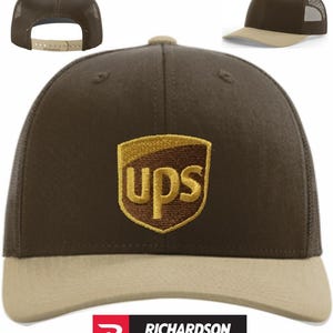 May include: A brown and tan trucker hat with the UPS logo embroidered on the front. The hat features a mesh back, a curved brim, and an adjustable snap closure. The Richardson brand logo is visible at the bottom.