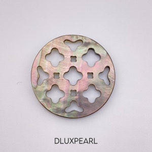 Puede incluir: Una incrustación redonda de nácar iridiscente con un patrón geométrico de recortes. La incrustación es de color rosa, verde y negro.
