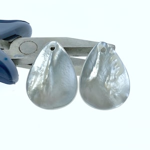 Può includere: Due ciondoli a forma di lacrima di colore bianco perlato con un foro nella parte superiore per la corda. I ciondoli sono lisci e hanno un leggero bagliore iridescente.