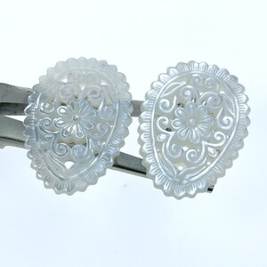 May include: A pair of white mother of pearl earrings with a delicate floral design. The earrings are oval shaped and have a scalloped edge.
