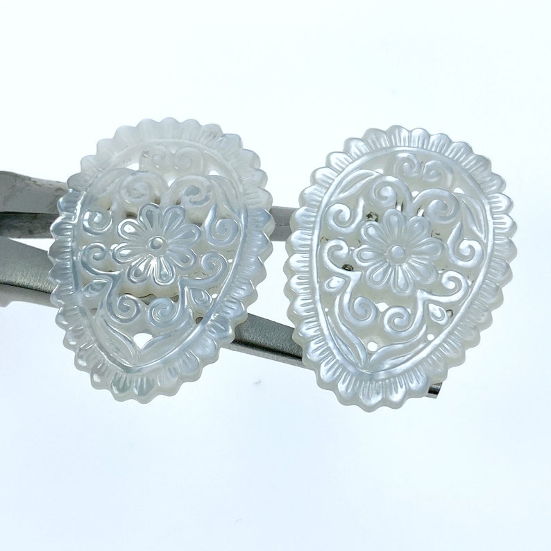 May include: A pair of white mother of pearl earrings with a delicate floral design. The earrings are oval shaped and have a scalloped edge.