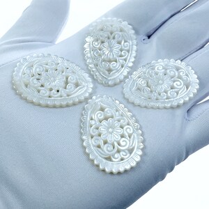 May include: Four white oval-shaped mother of pearl pendants with intricate floral carvings. The pendants are on a white surface.