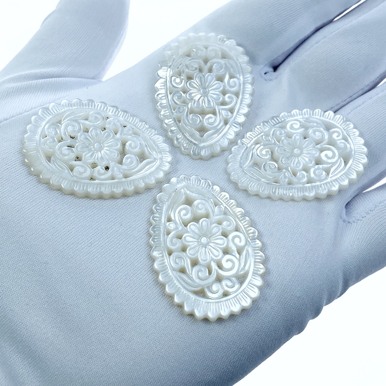 May include: Four white oval-shaped mother of pearl pendants with intricate floral carvings. The pendants are on a white surface.