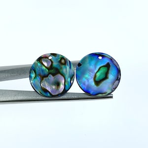 Puede incluir: Dos colgantes redondos de concha de abulón iridiscente con un agujero en el centro. Las conchas son azules, verdes y moradas con marcas negras.