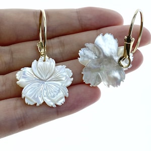 Peut inclure: Une paire de boucles d'oreilles créoles dorées avec des breloques de fleurs en nacre blanche. Les fleurs ont un design sculpté et une finition brillante.