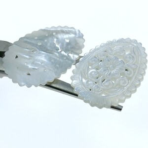 May include: Two white, intricately carved mother of pearl shell embellishments. One is oval shaped with a floral design, the other is a more simple oval with a repeating pattern.
