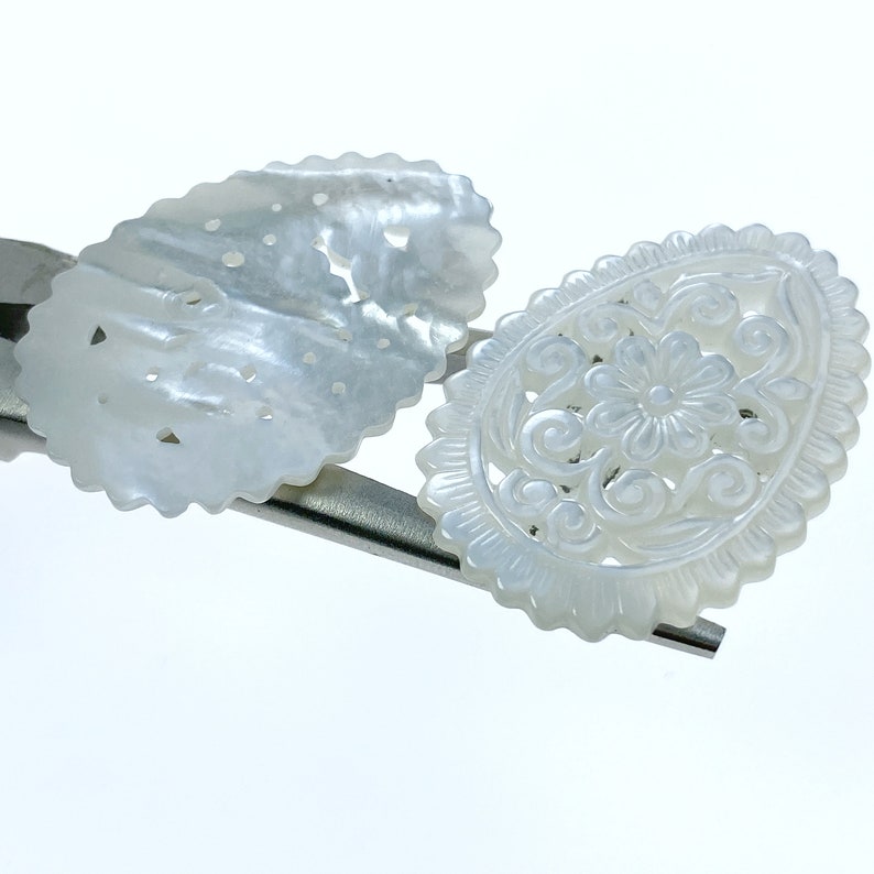 May include: Two white, intricately carved mother of pearl shell embellishments. One is oval shaped with a floral design, the other is a more simple oval with a repeating pattern.
