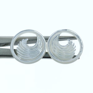 May include: A pair of white mother of pearl earrings with a wave design. The earrings are round and have a silver tone.