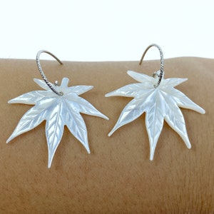 May include: A pair of white mother of pearl earrings shaped like maple leaves. The earrings are silver hoop style.